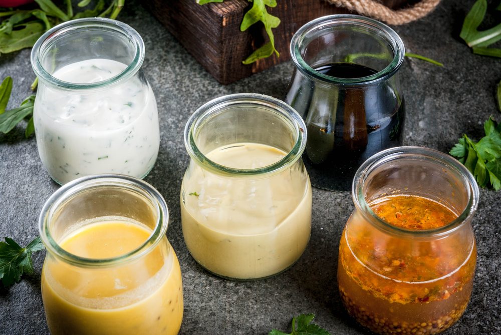 Set of dressings for salad: sauce vinaigrette, mustard, mayonnaise or ranch, balsamic or soy, basil with yogurt. Dark stone table. On background of greenery, vegetables for salad. Copy space