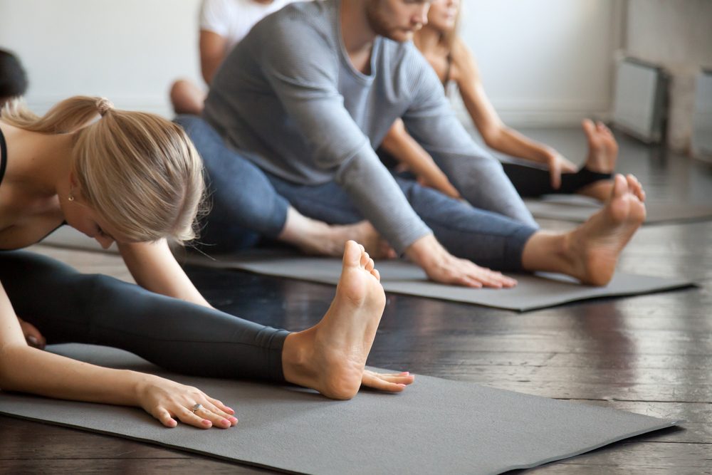 Young sporty people practicing yoga lesson with instructor, sitting in Janu Sirsasana exercise, Head to Knee Forward Bend pose, working out, indoor close up image, studio. Wellbeing, wellness concept 