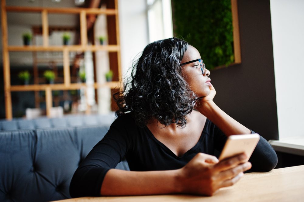 African american girl in sunglasses sitting at cafe and holding mobile phone at hand.