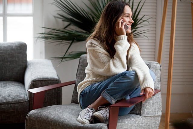 Happy cheerful young woman talking on the phone at home, smiling teen girl making answering call by cellphone sitting on chair, beautiful lady having pleasant funny conversation speaking by mobile