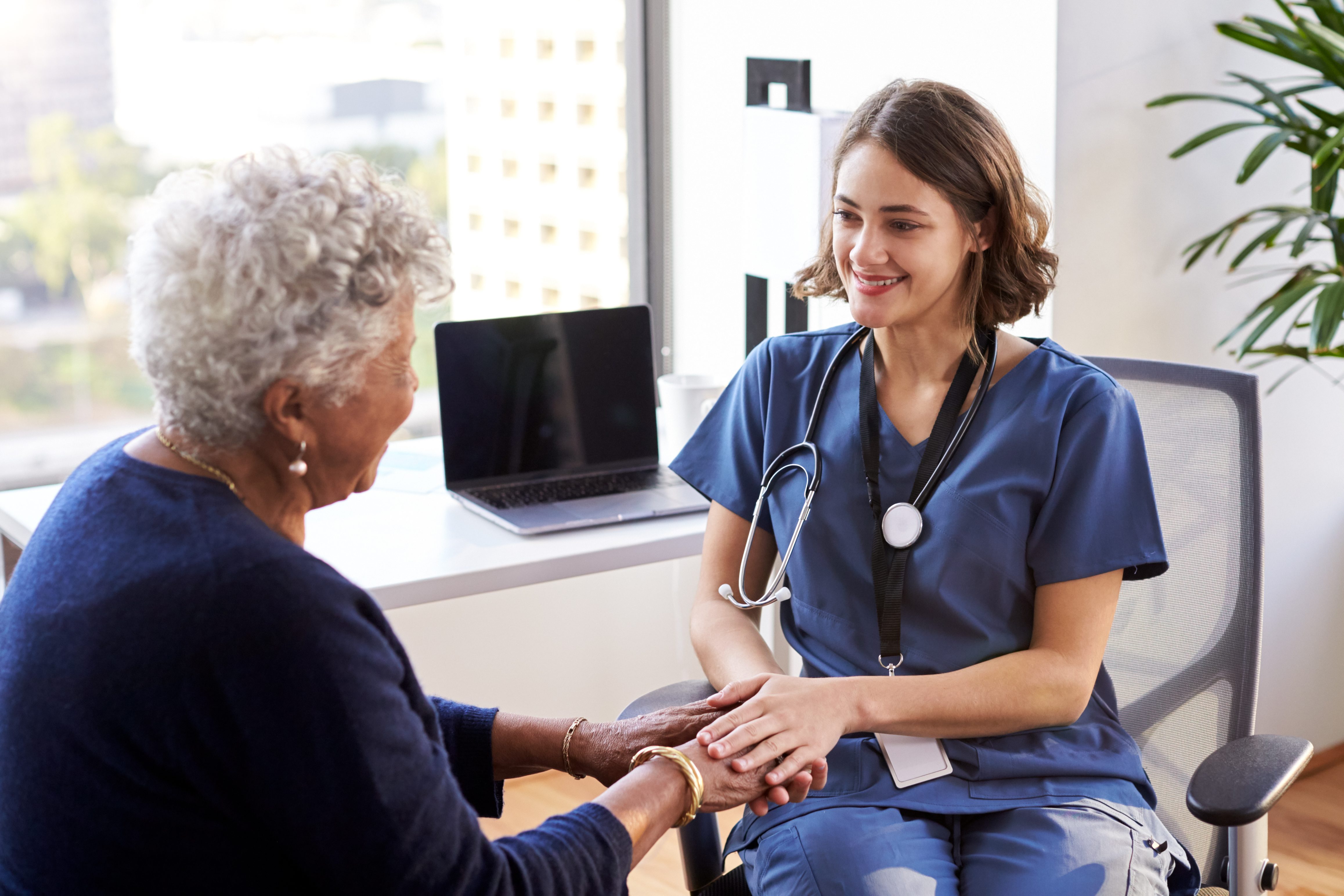 Nurse Wearing Scrubs In Office Reassuring Senior Female Patient And Holding Her Hands