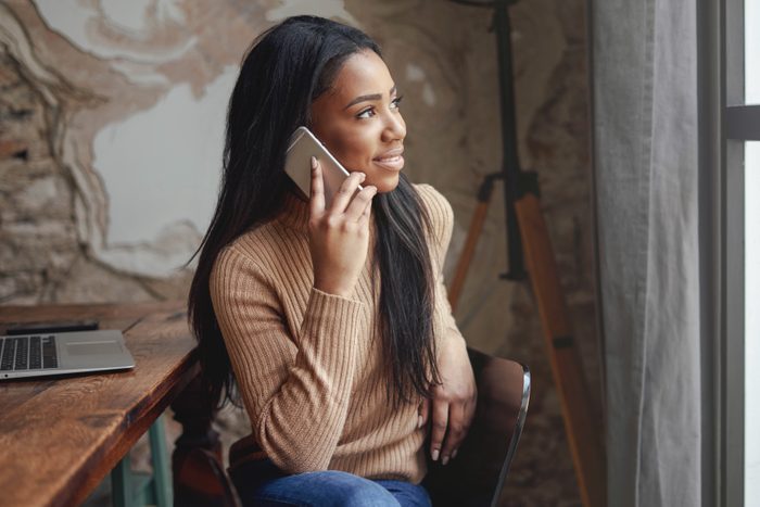 Beautiful african woman is talking on a mobile phone while sitting in the office with a laptop. Young freelancer girl is calling to the clients and reading emails on a portable computer.