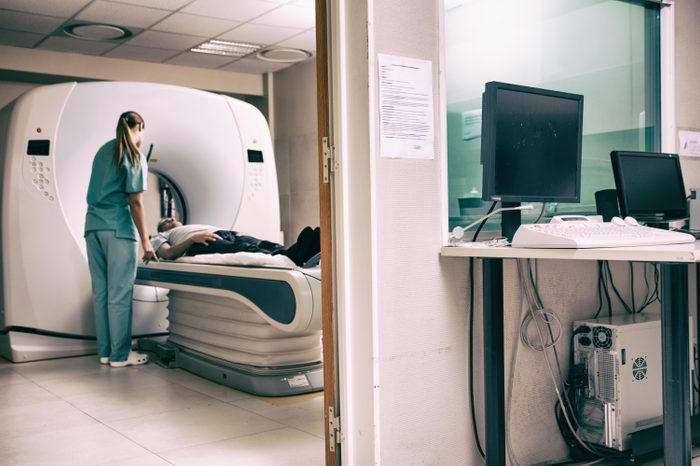 Hospital room with computer and monitors. Female doctor and male patient undergoing scan in adiacent room.