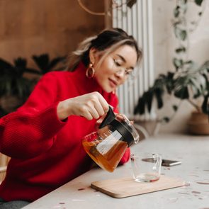 Snapshot of Asian girl in red outfit pouring herself green tea in cup while sitting in cafe