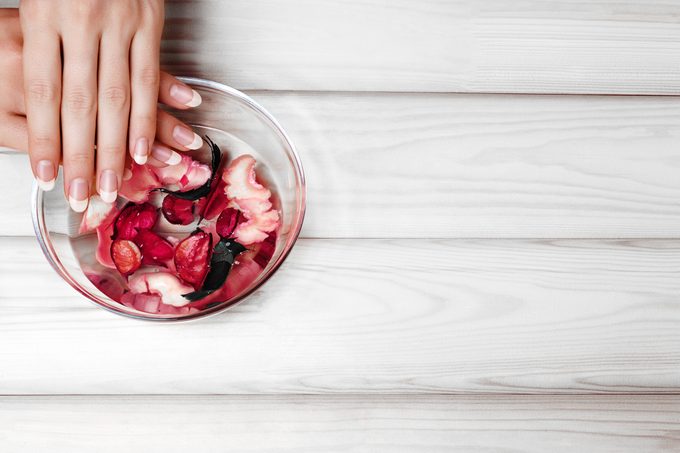Gentle Natural Manicure, Hand Care. Soaking Finger Nails in Bath with Herbal Oil and Floral on White Wooden Table with Copy Space