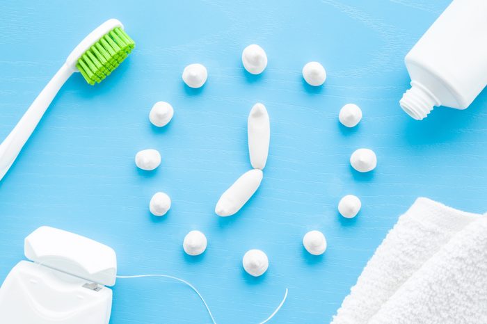 Toothbrush, white tube of toothpaste, dental floss and towel on pastel blue background. Clock created from paste. Healthy teeth. Morning routine concept. 