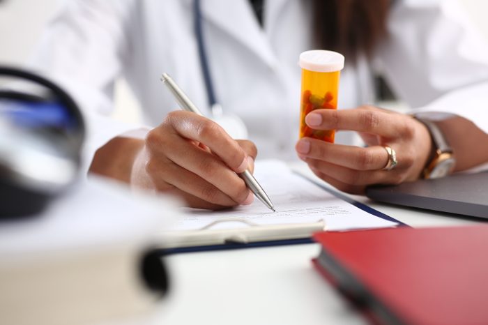 Female black medicine doctor hand hold jar of pills and write prescription to patient at worktable. Panacea and life save, prescribing treatment legal drug store concept. Empty form ready to be used