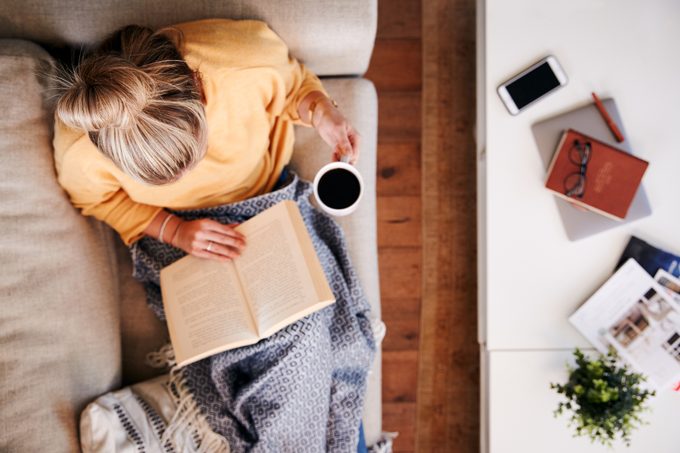 woman at home sitting on couch reading a book and drinking coffee