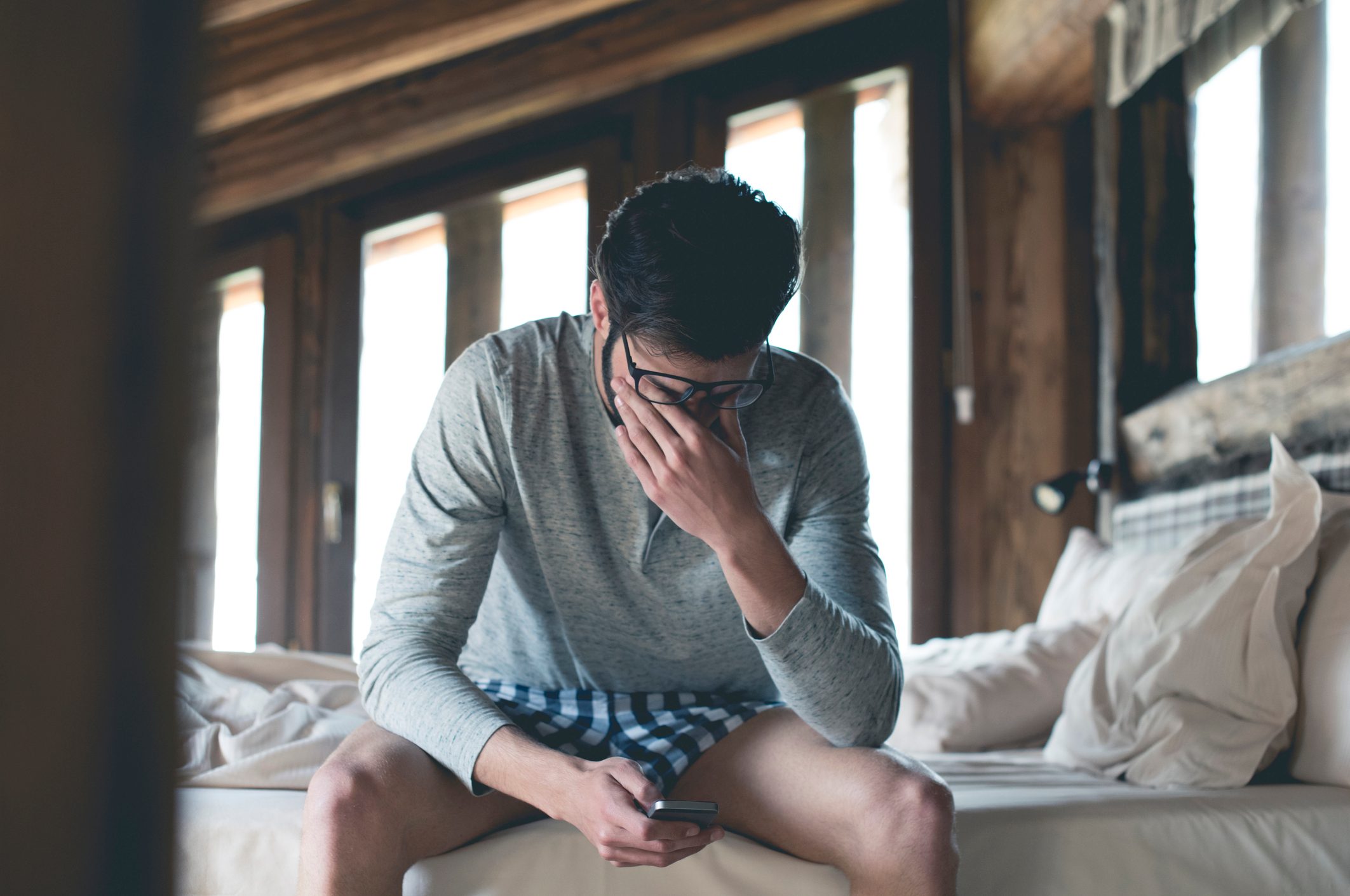 tired young man sitting on bed looking at smartphone