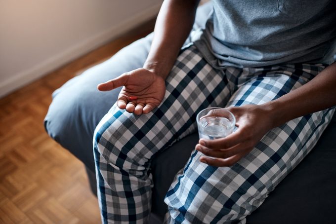 cropped shot of a man sitting on bed with glass of water and painkiller in hands