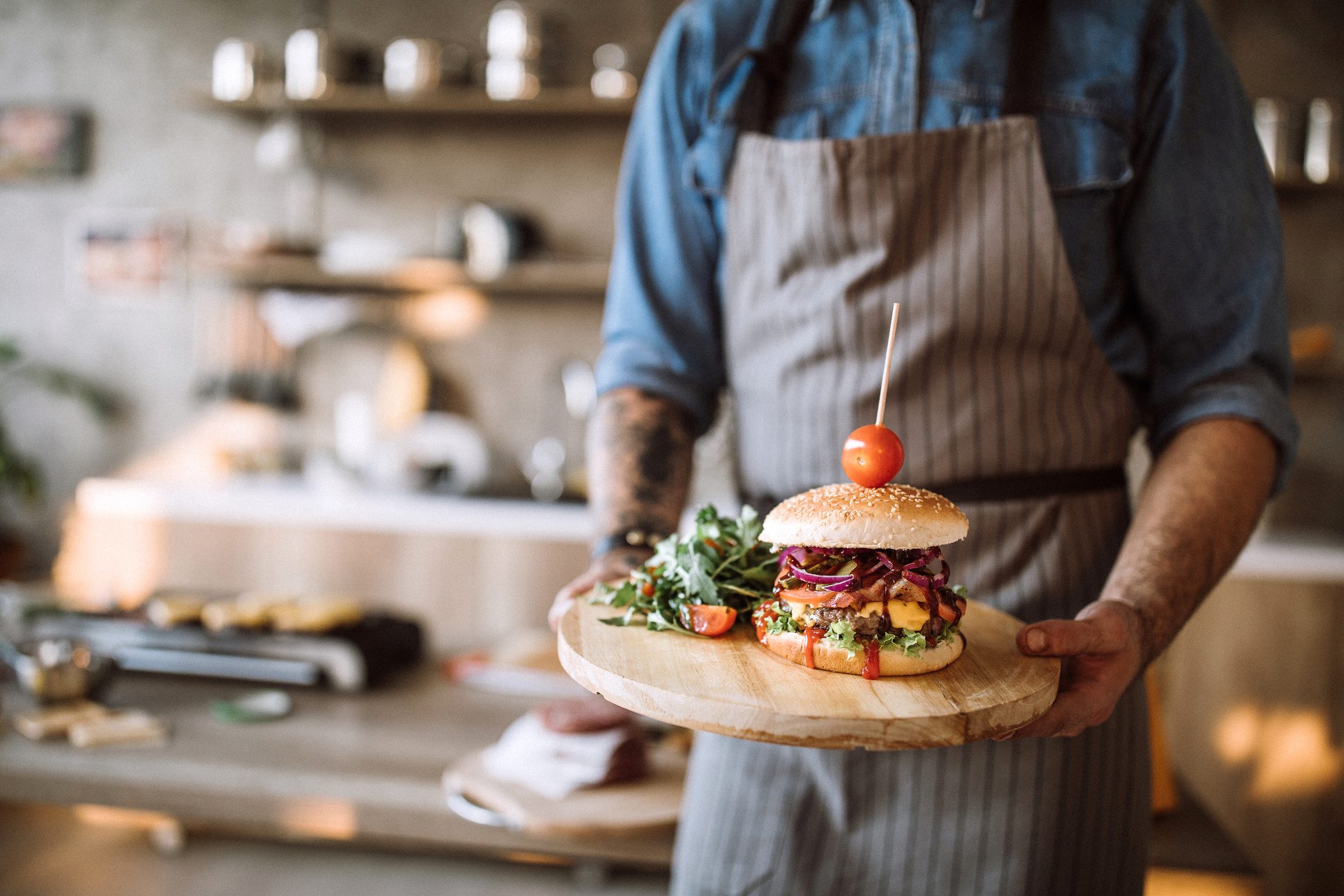 Man with apron standing in the kitchen and holding a wooden plate with prepared delicious hamburgers