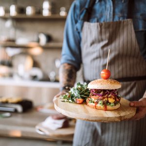 Man with apron standing in the kitchen and holding a wooden plate with prepared delicious hamburgers