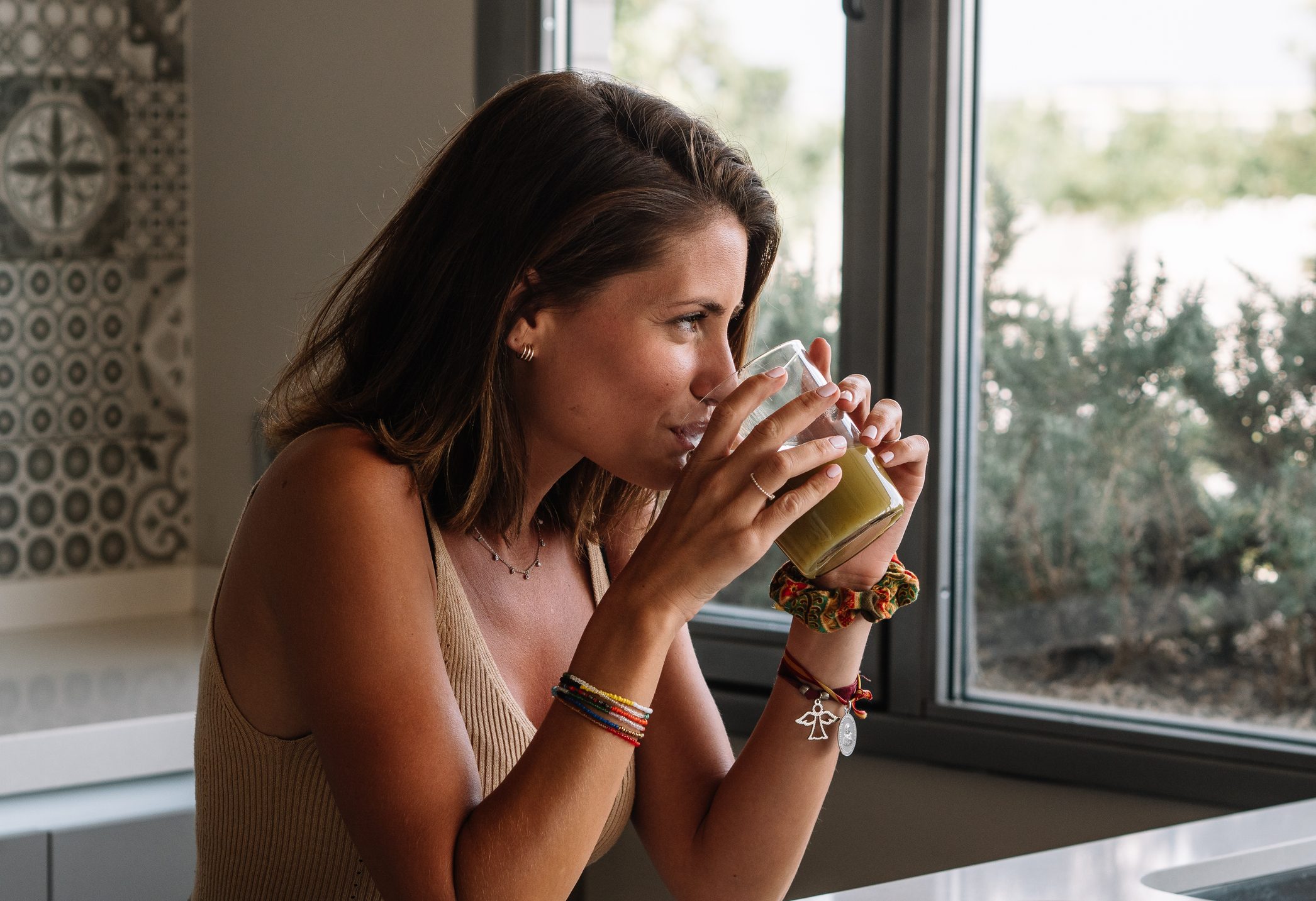 Caucasian girl is having a green vegetable juice in front of her big kitchen window.