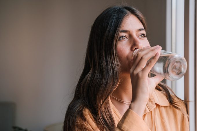 Young Woman Drinking Water In Glass Bottle By Window