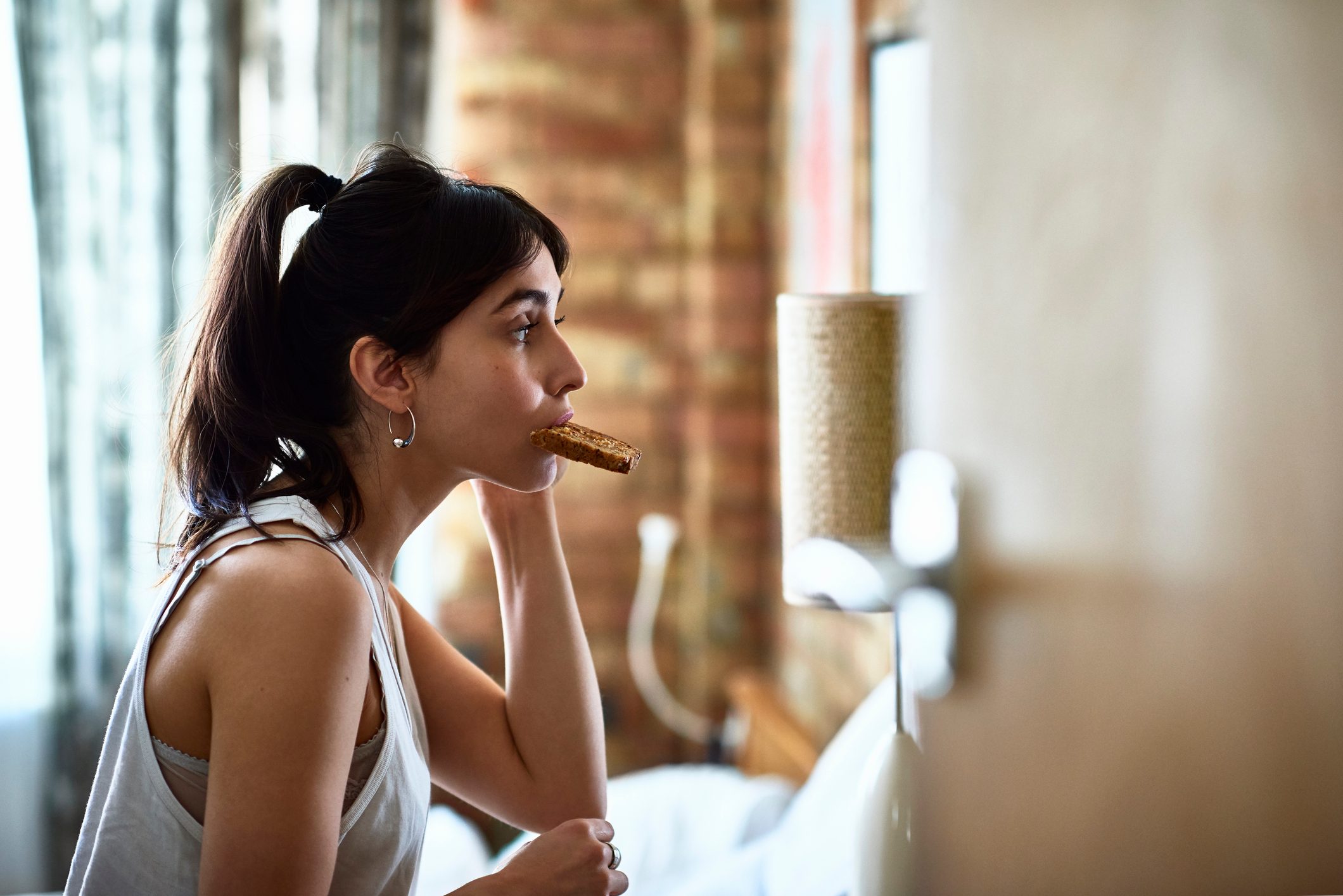 Young woman biting piece of toast and checking herself in mirror