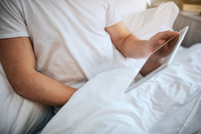 young man in bed with hand under blanket and tablet in hand