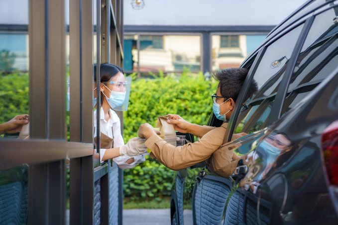 man in protective mask taking food bag and coffee with woman waitress wearing face mask and face shield at drive thru during coronavirus outbreak.