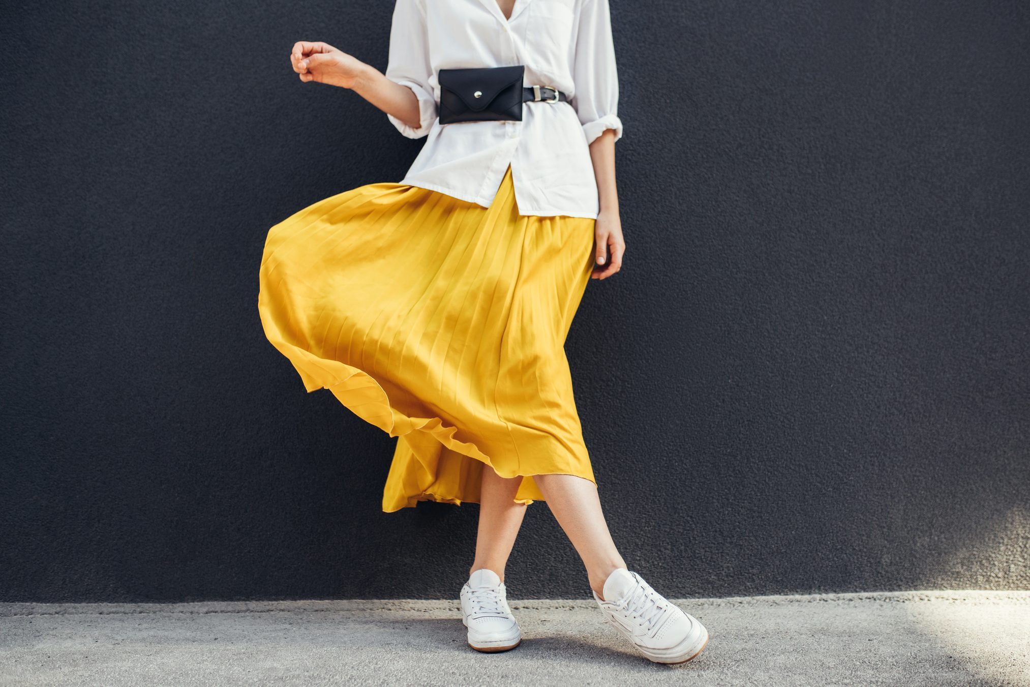 Horizontal cropped image of stylish slim woman in beautiful yellow skirt. Caucasian female fashion model standing over gray wall background outdoor with copy space.