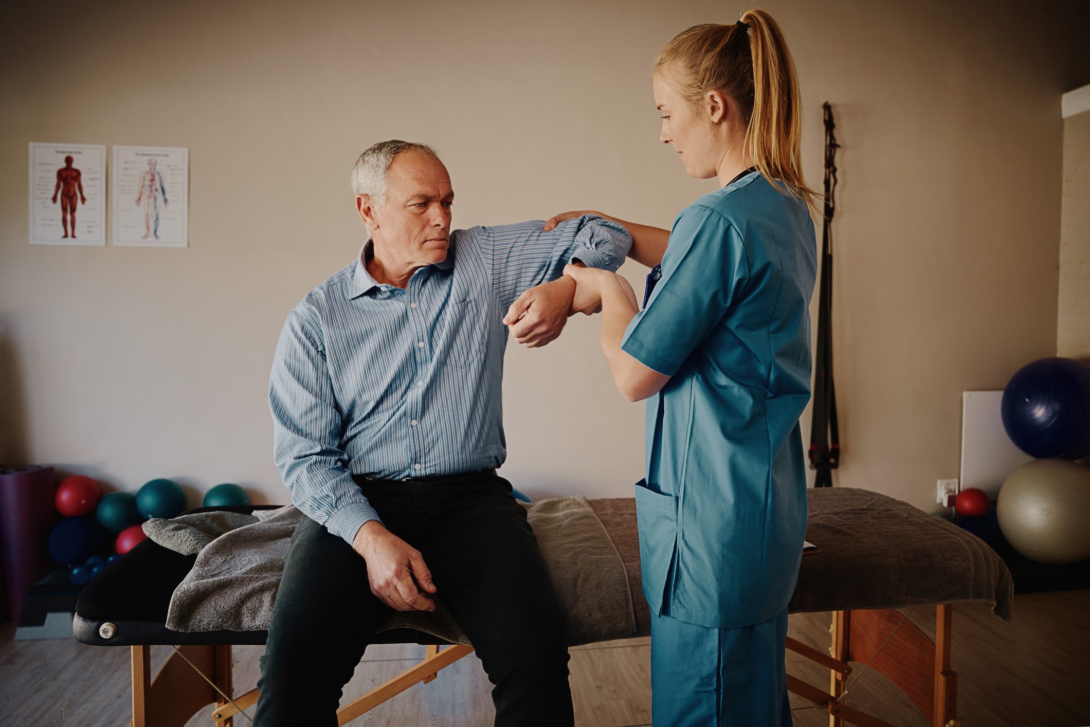 Young female physiotherapist helping senior man with elbow exercise in clinic - young female doctor helping senior patient