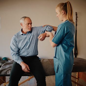 Young female physiotherapist helping senior man with elbow exercise in clinic - young female doctor helping senior patient