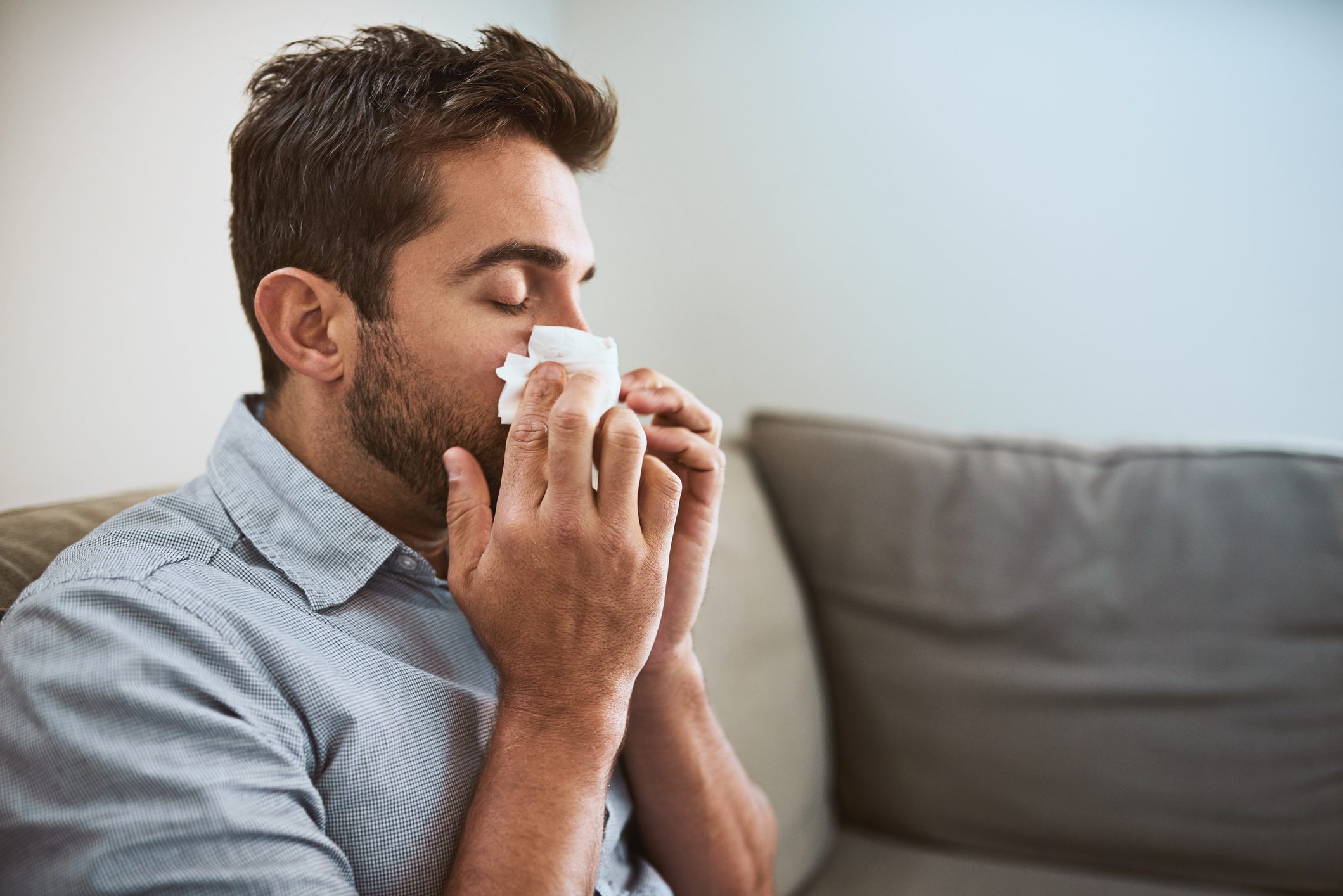 close up of young man blowing his nose while sitting on couch at home