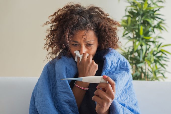 woman sick at home, wrapped in a blanket, holding a tissue to her nose with one hand and a thermometer in the other wondering if it's the flu, covid, or flurona