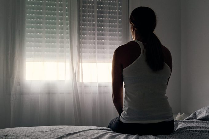Rear view of an unrecognizable abused woman sitting on her bed looking out the window.