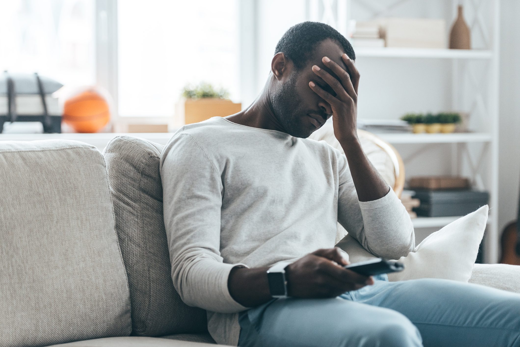 man on couch in living room feeling bad about what he's seeing on the news. he has the tv remote in one hand, the other hand is on his face to indicate stress.