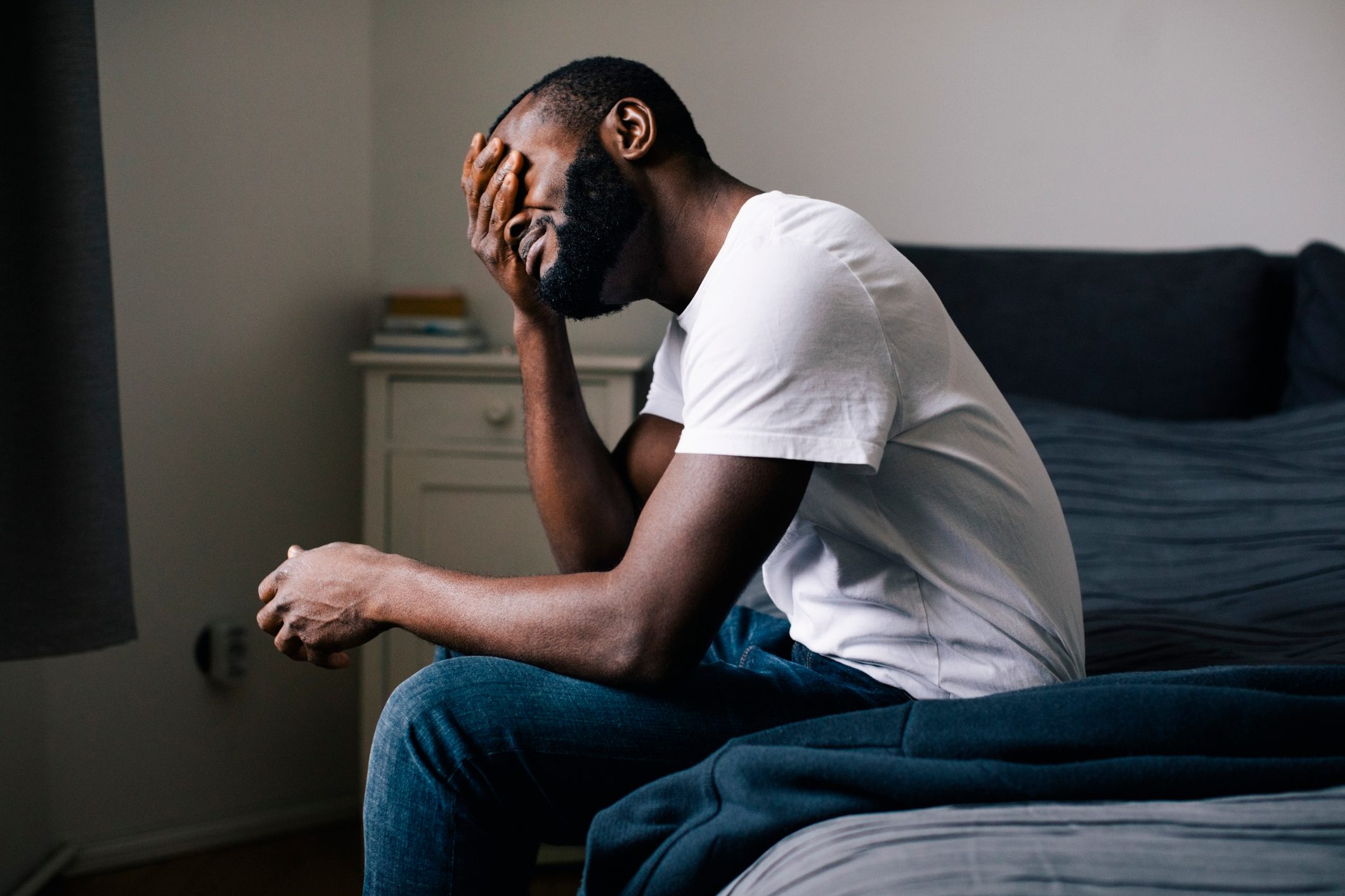 Emotionally stressed man covering eyes with hand sitting on bed at home