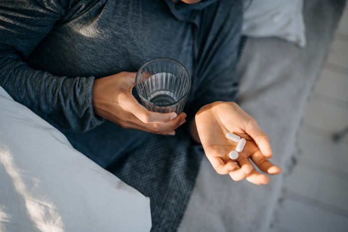 Cropped shot of young Asian woman lying in bed and feeling sick, taking medicines in hand with a glass of water