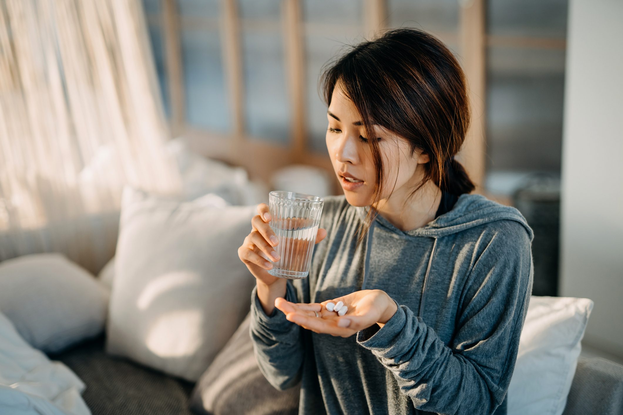 Young Asian woman sitting on bed and feeling sick, taking medicines in hand with a glass of water