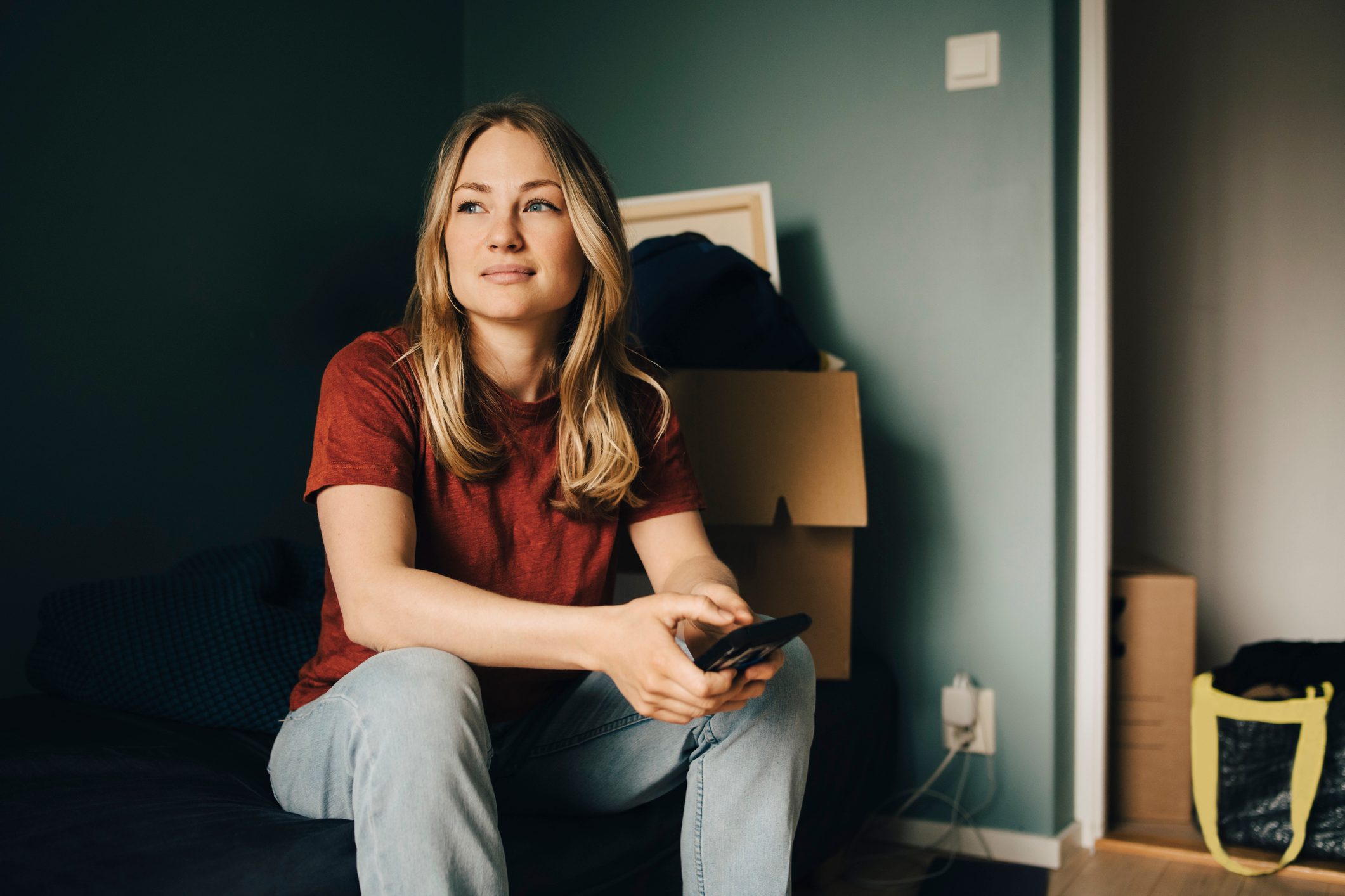 Thoughtful young woman looking away while sitting with smart phone in bedroom