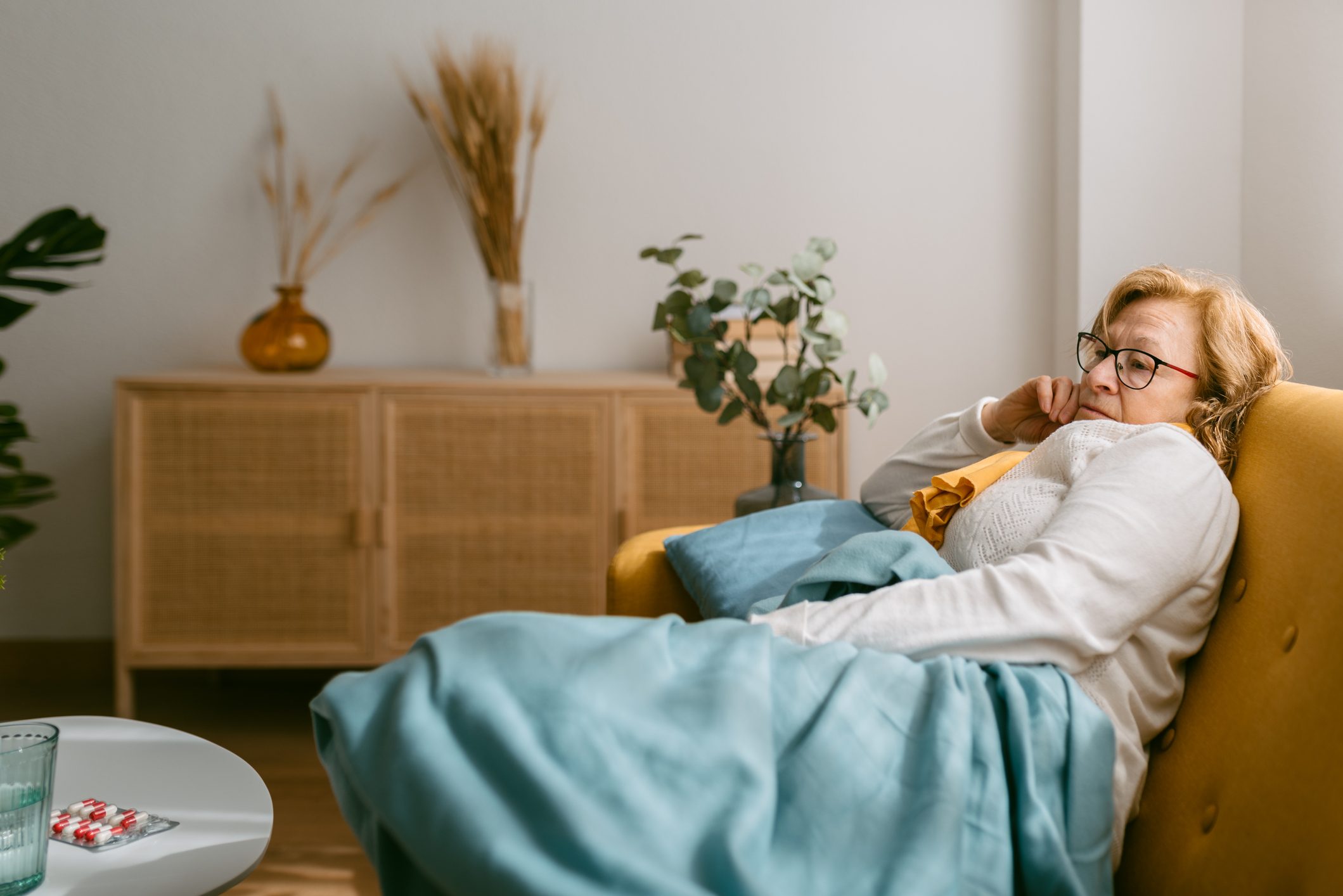 Caucasian woman with blonde hair, sick lying on the sofa in the living room of her house while covering herself with a blue blanket