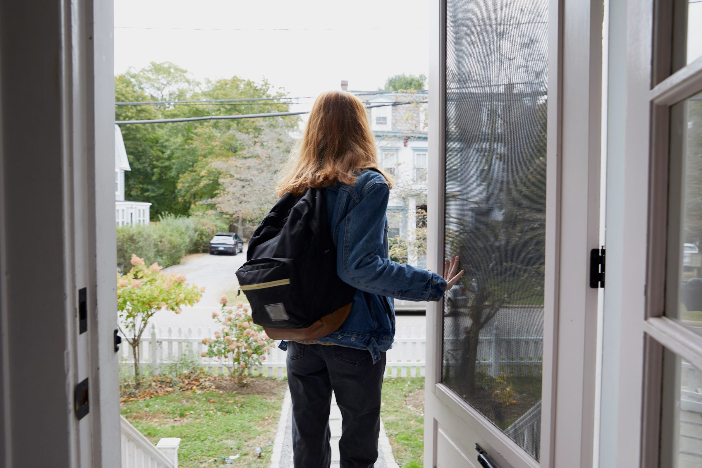 Teenage girl walking out the front door of her house. Back view of her leaving the house. She is on her way to school, wearing a back pack and holding the door open.