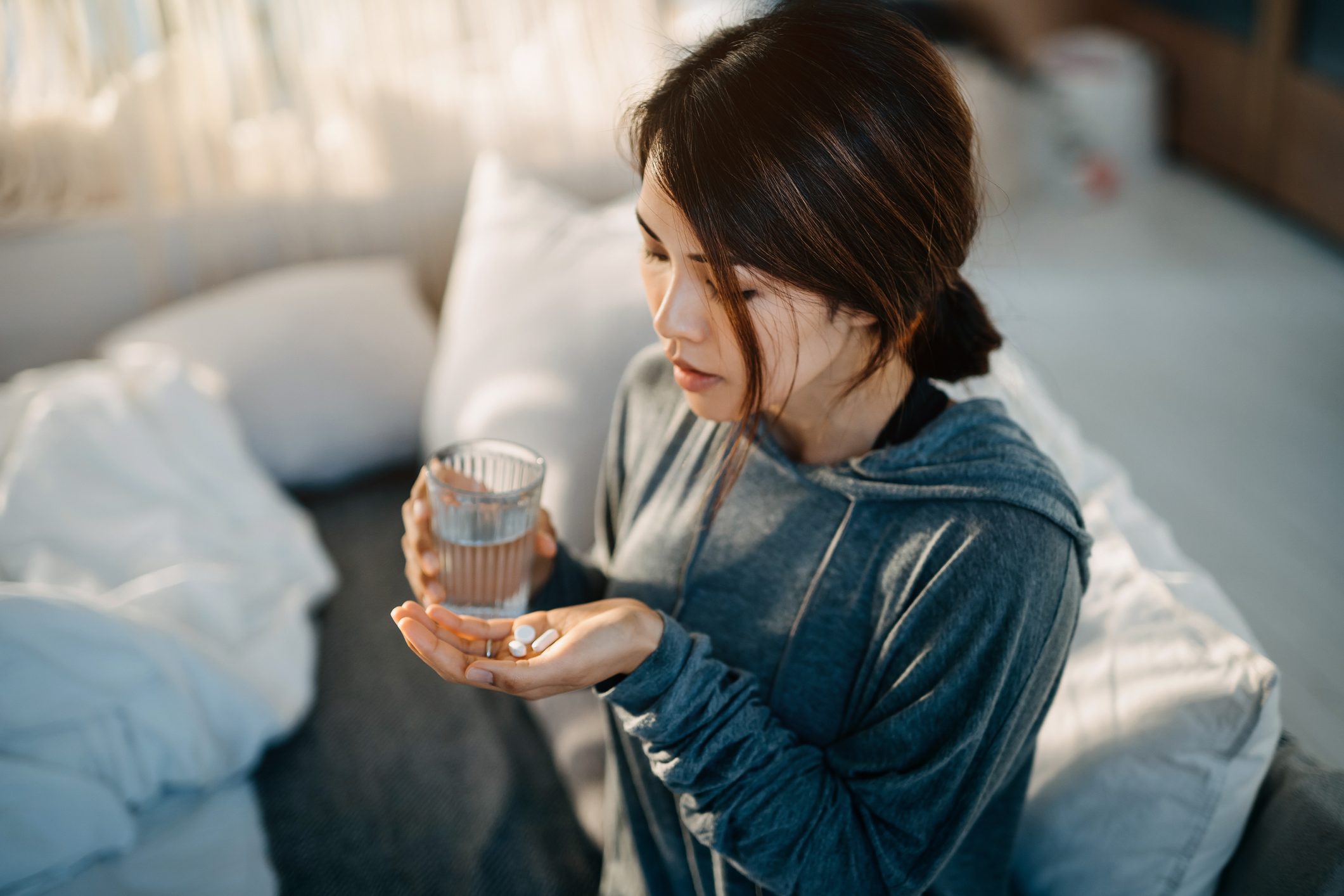 Young Asian woman sitting on bed and feeling sick, taking medicines in hand with a glass of water