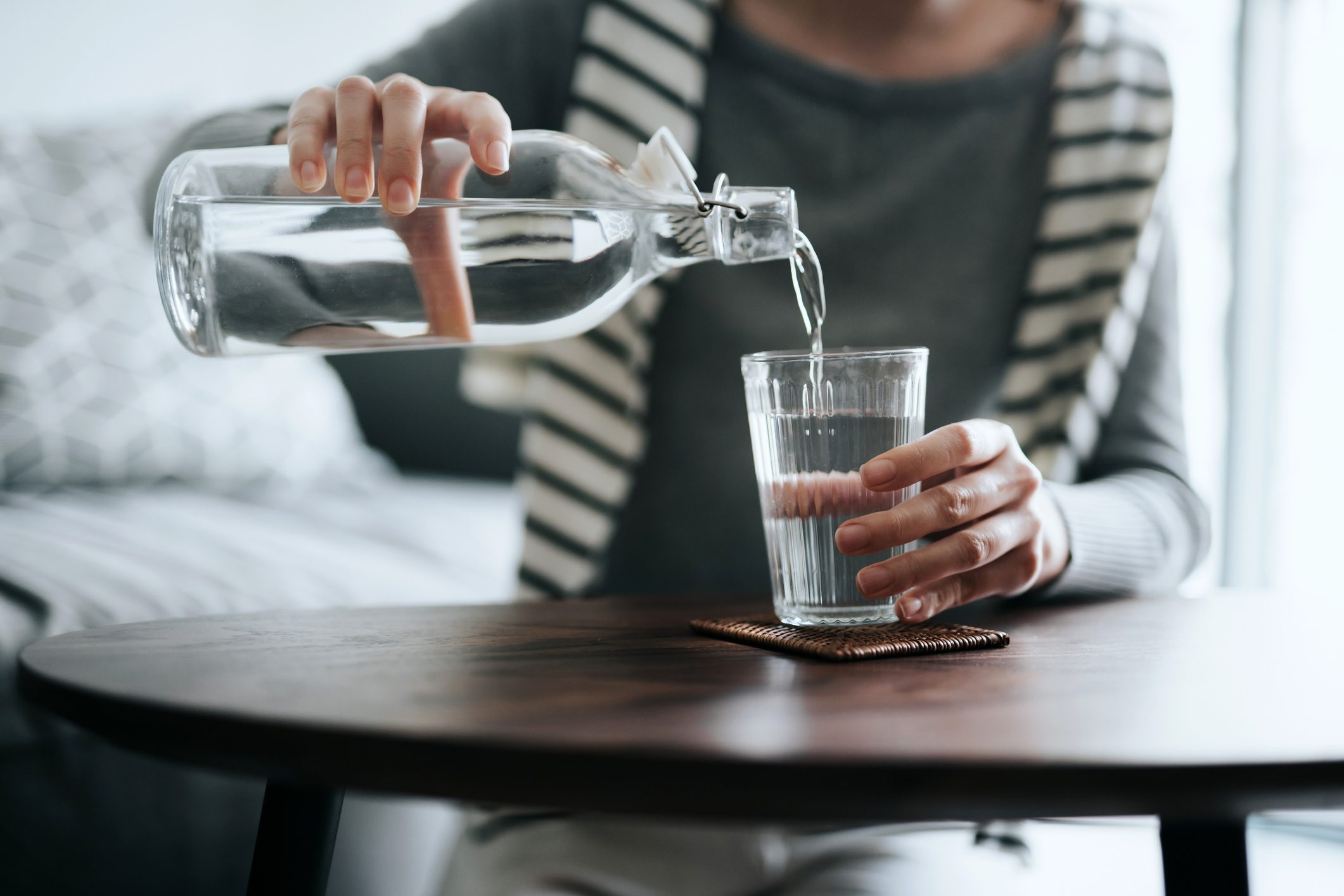 Close up of young Asian woman pouring water from bottle into the glass on a coffee table at home. Healthy lifestyle and stay hydrated