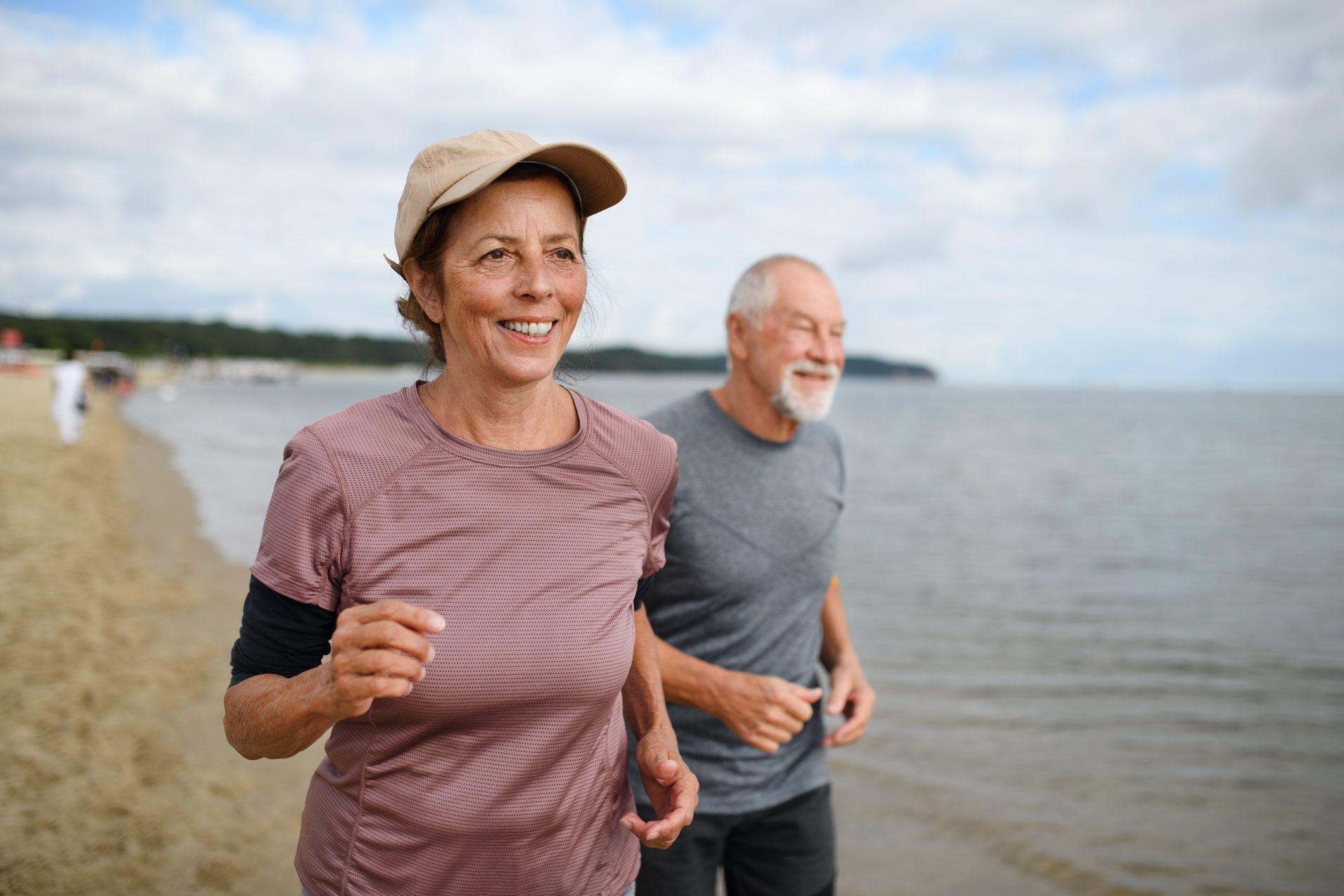 Active senior couple runners jogging outdoors on sandy beach by sea in early morning.