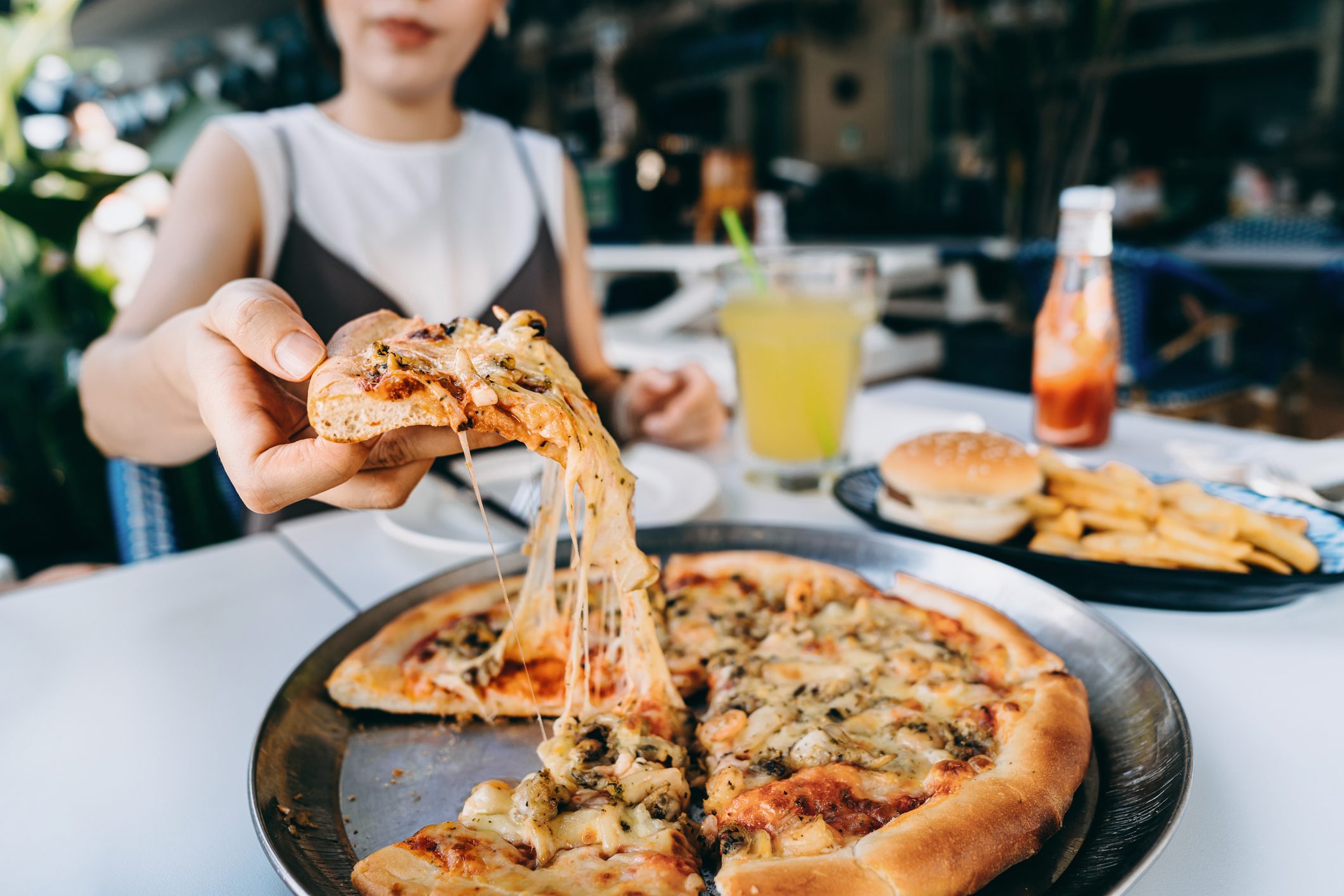 Close up of young Asian woman getting a slice of freshly made cheesy seafood pizza, enjoying her lunch in an outdoor restaurant. Eating out lifestyle