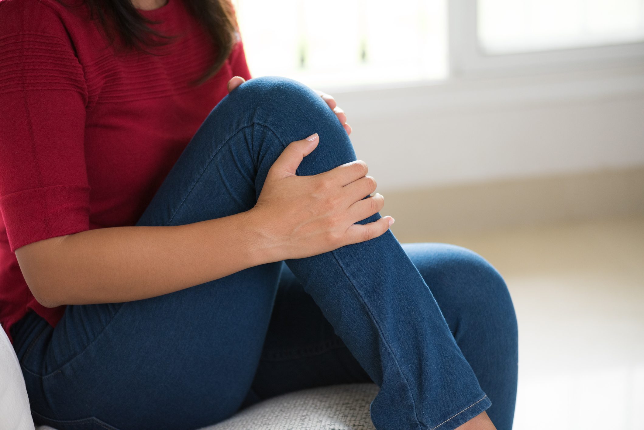 Closeup young woman sitting on sofa and feeling knee pain and she massage her knee at home. Healthcare and medical concept.