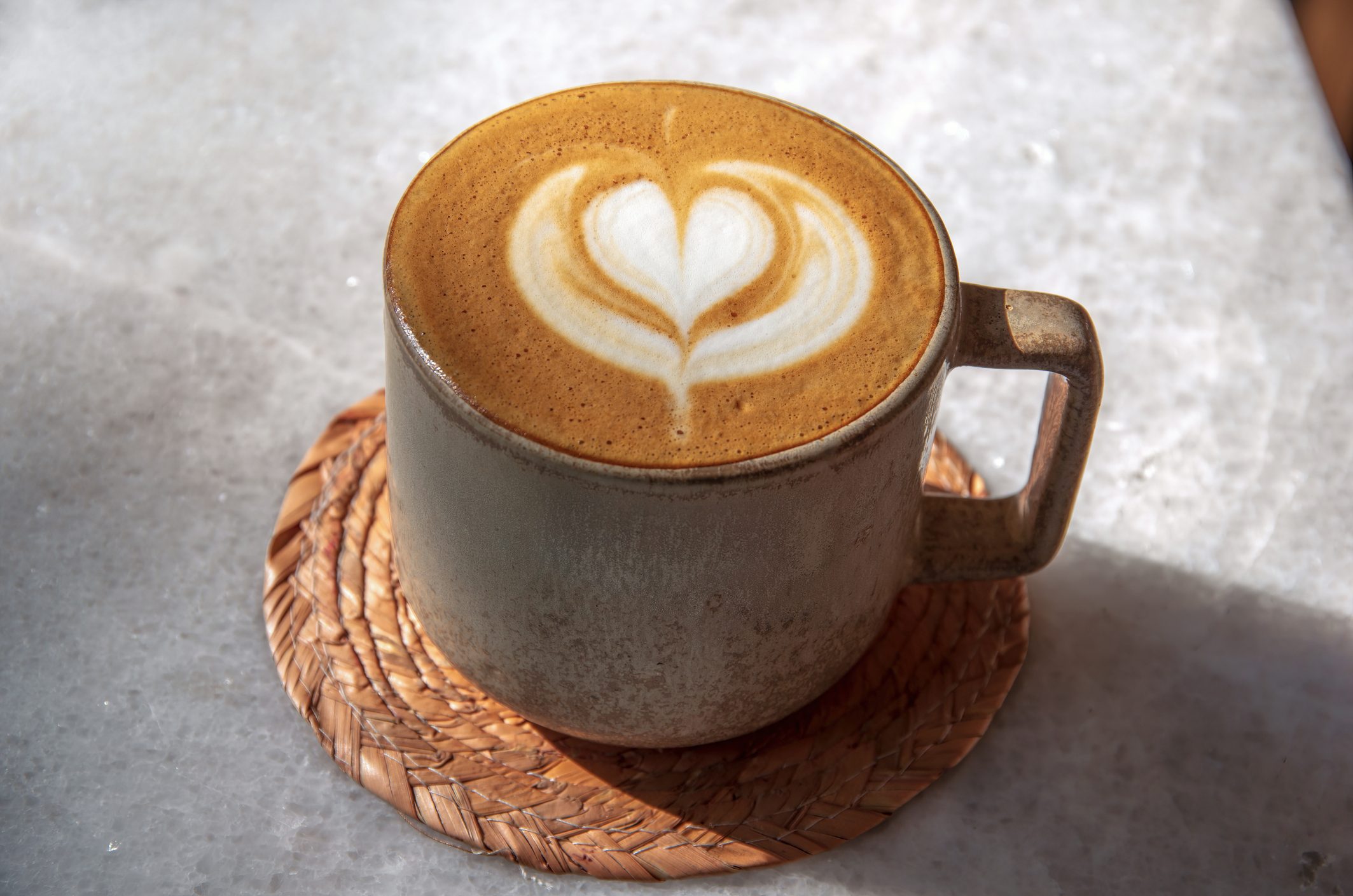 Flat white coffee in a gray ceramic cup on a plaited straw coater on a quartz topped outdoor cafe table