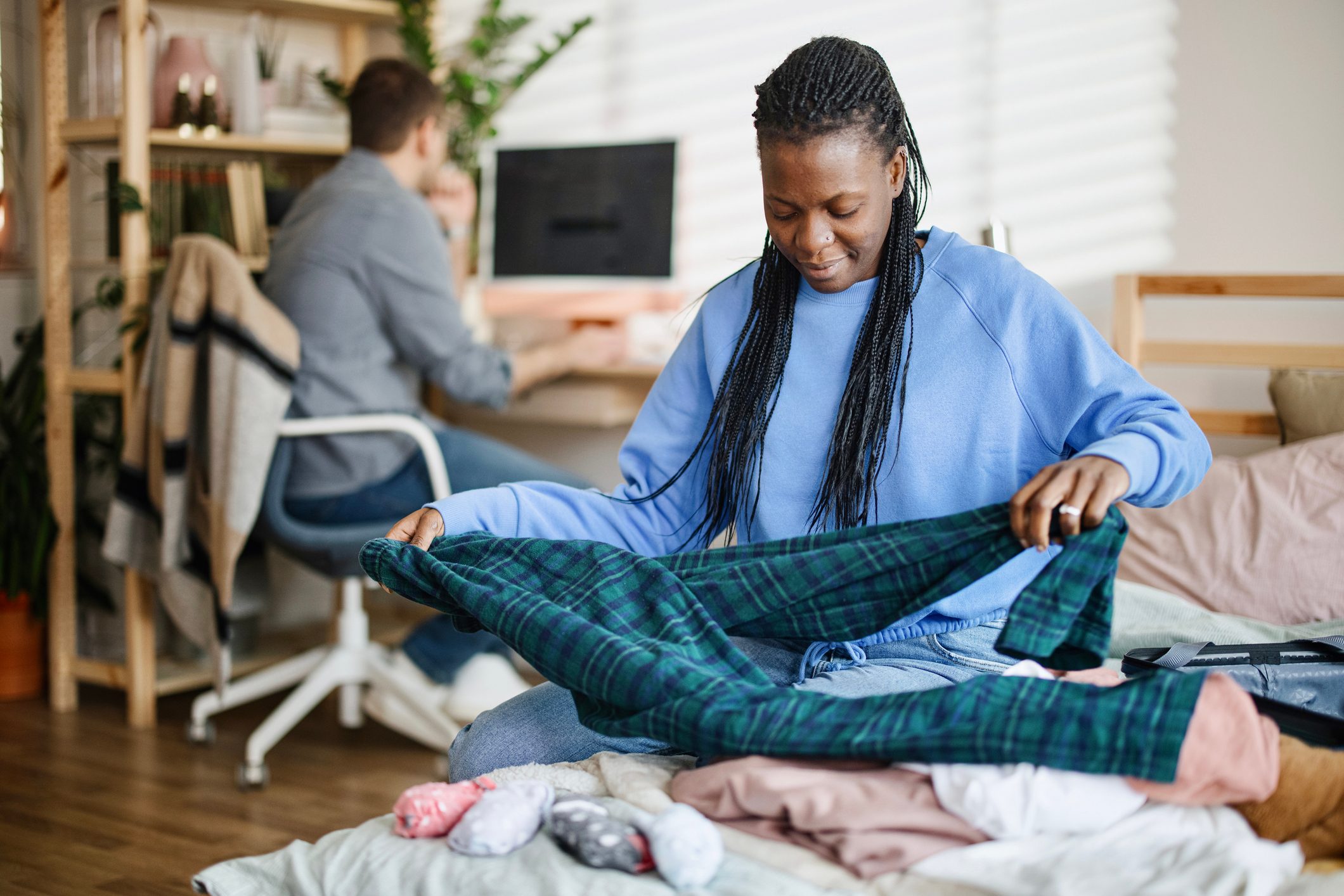 A woman folds her pyjamas while her husband sits in the background and looks at a desktop computer