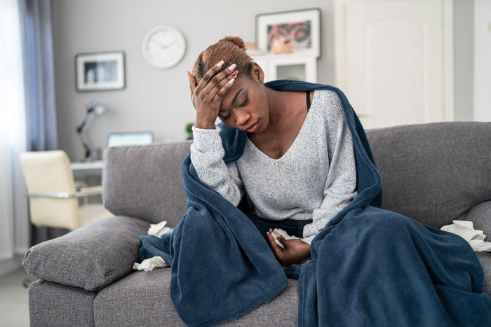 Young african american woman, sitting on the sofa at home, having a fever and touching her forehead to check her temperature