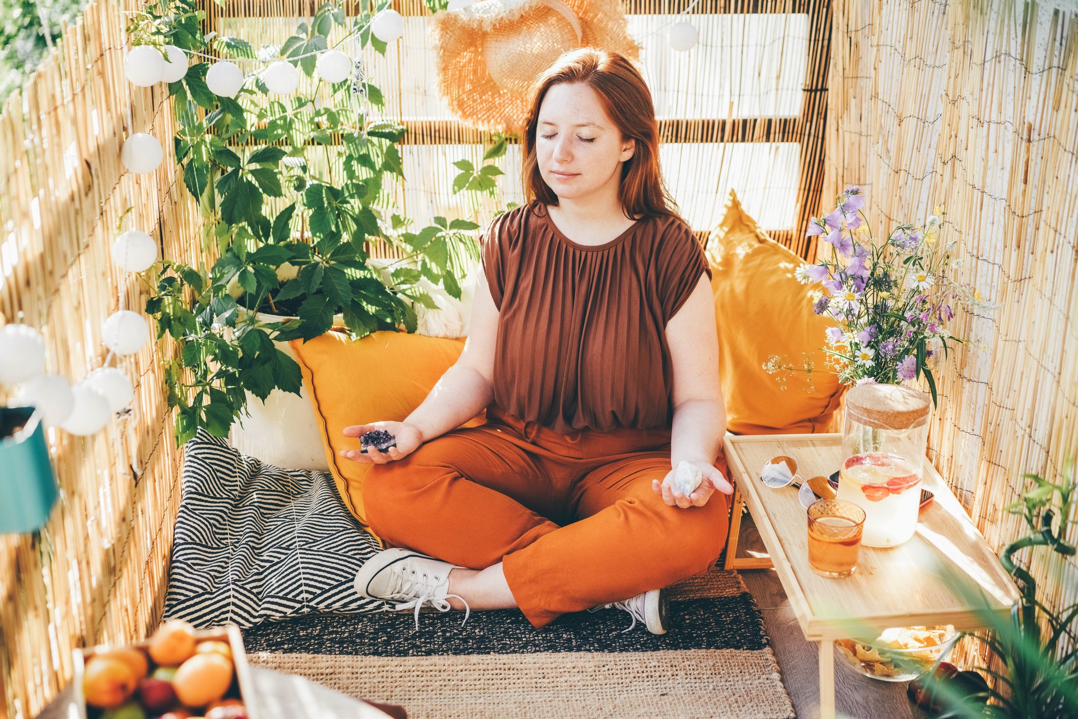 Young woman meditating in lotus pose with shiny crystals on decorated sunny terrace.