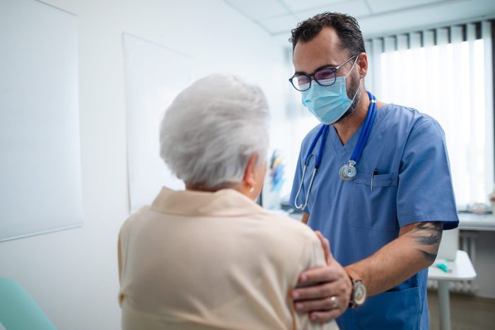 Rear view of senior woman with doctor wearing protective face masks and talking warmly in doctor's office.