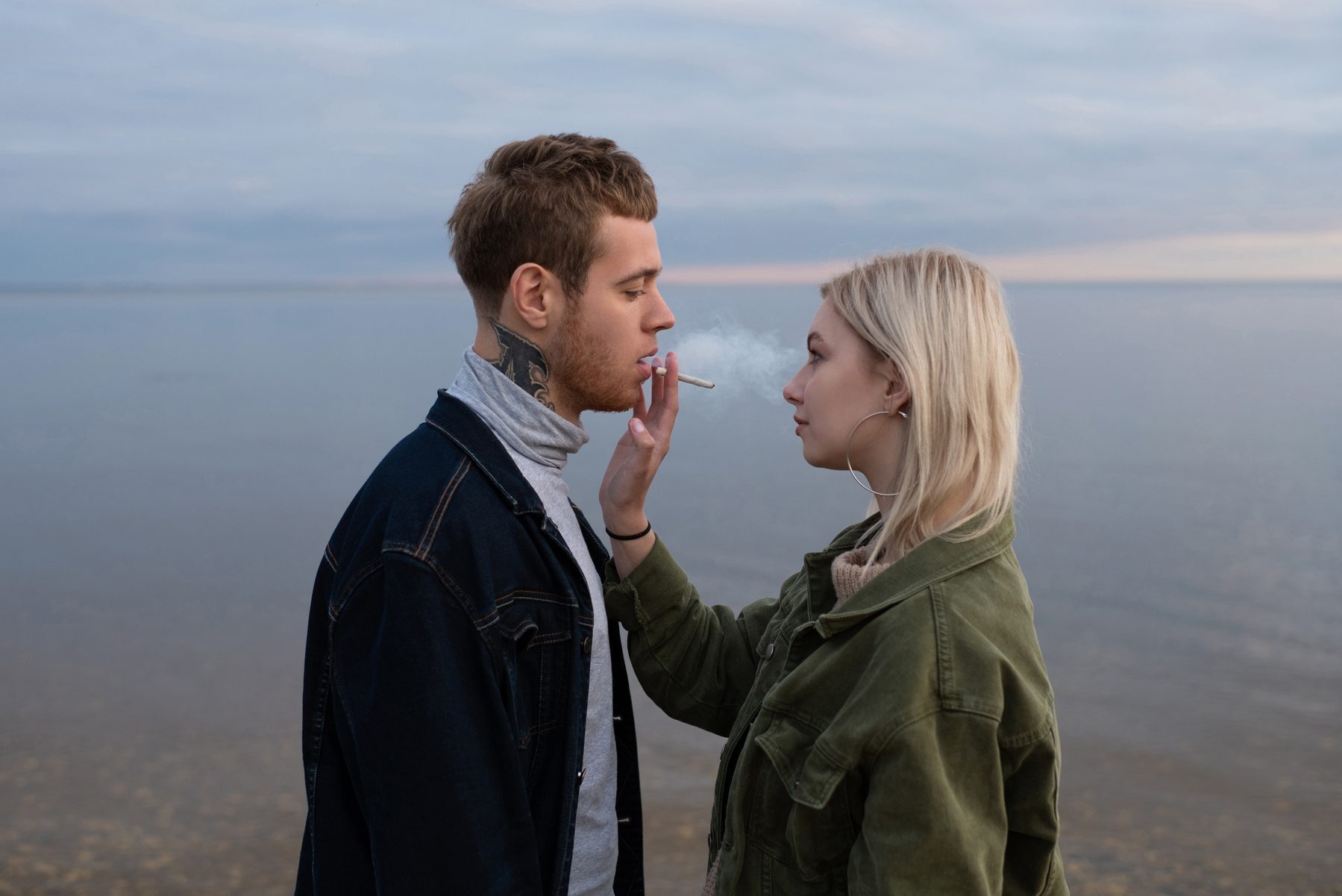 Sensual young hipsters sharing joint on dusky coast