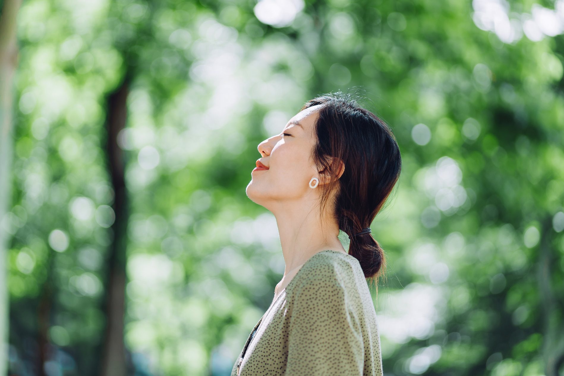 Beautiful young Asian woman meditating in the nature with her eyes closed, setting herself free and feeling relieved. Enjoying fresh air and breathing in the calmness with head up against sunlight in the morning. Freedom in nature. Connection with nature