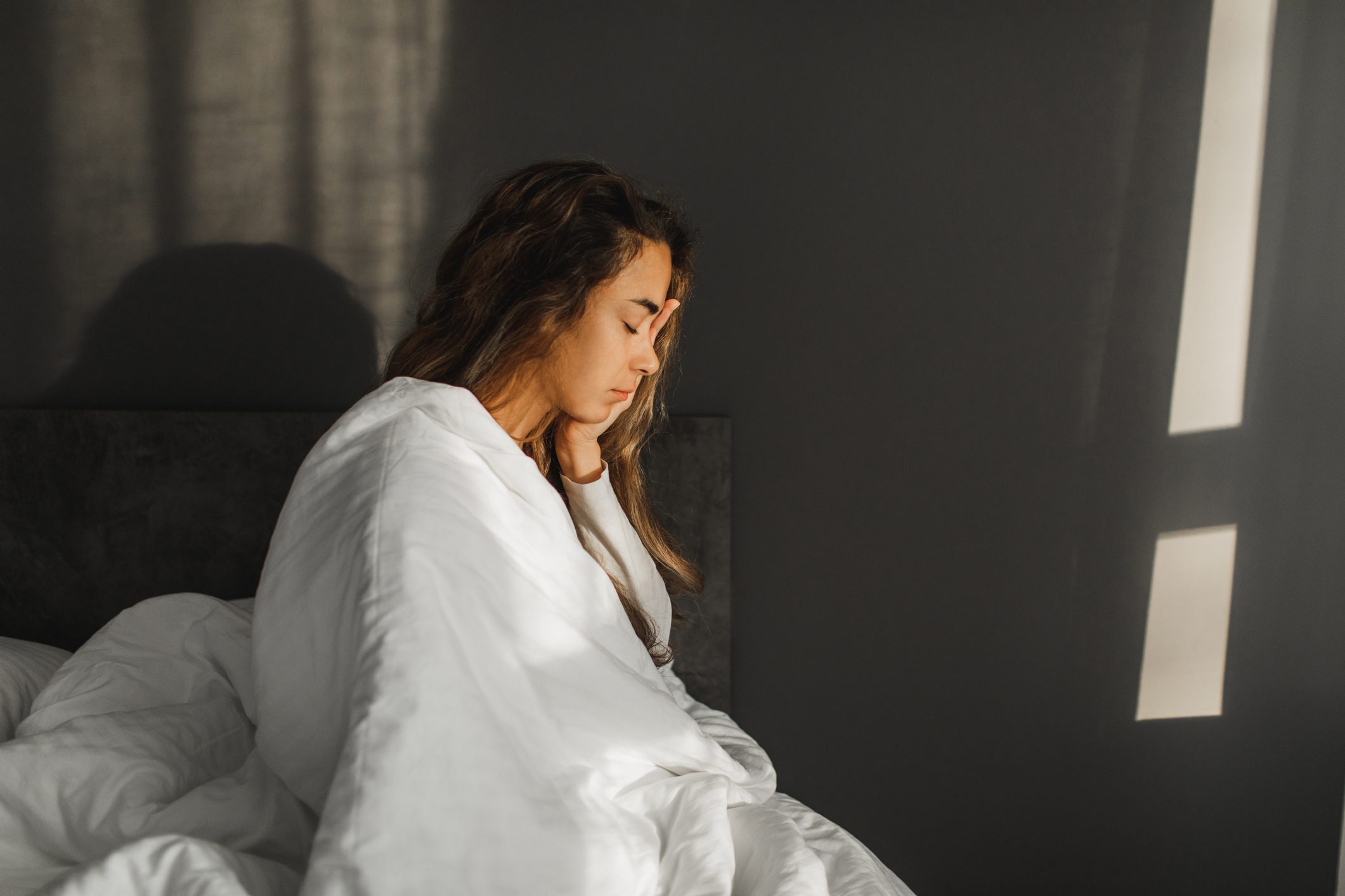 Woman yawning and can't wake up in morning. Sitting in bed covered with blanket.