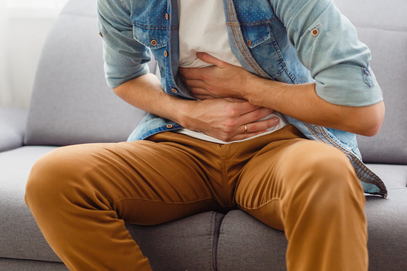 The man is sitting on a gray couch and holding his belly. Medicine and health concept, stomach problems. The man suffers from stomach ache, gastric problems. Abdominal pain, suffering and pain.