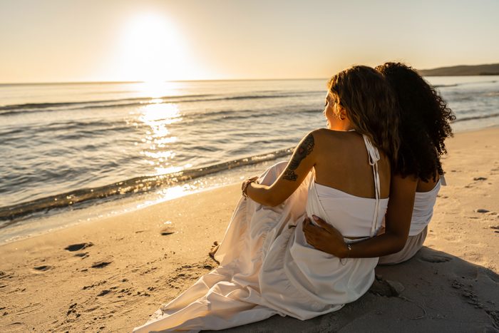 Two lesbian young women view from back sitting on seashore looking at sunset or dawn. Afro american homosexual girl hugs her blonde girlfriend near ocean water in summer vacations. Diversity concept