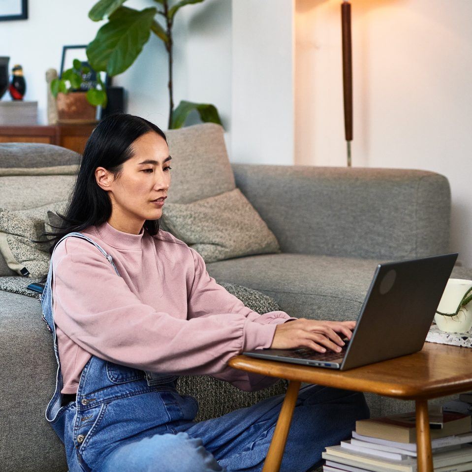 Mature Chinese woman sitting on living room floor home using laptop, working from home